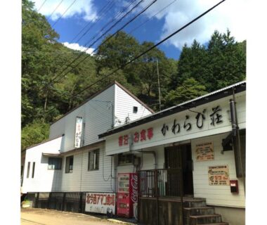 Exterior of white minshuku with Japanese signage, vending machine, steps and forested mountain backdrop in Nara, Kansai