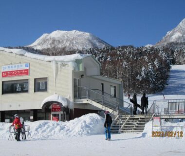 Snow-covered building and ski slope at Minshuku Mutsukari in Hokkaidō with skiers and mountain backdrop