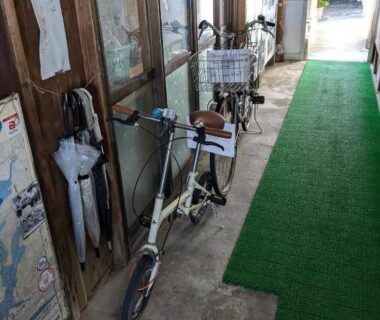 Corridor with parked bicycles and umbrellas beside wooden panels and green carpet runner at Minshuku Takahashi in Shizuoka