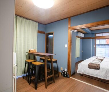 Western-style room with bed, small wooden dining table and stools, green curtains and folded towels at Mitomi Inn Tokyo