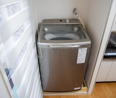 Silver top-loading washing machine beside kitchenette and window blinds in a compact Mitomi inn room, Tokyo, Kantō