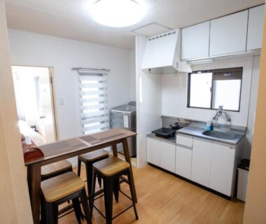 Compact kitchenette with wooden bar counter and three stools, adjacent sleeping area at Mitomi inn, Tokyo