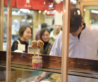 Vendor arranging skewered snacks behind glass counter with customers, Miyajima Morinoyado, Chūgoku, Hiroshima, Japan