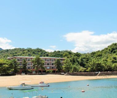 Sandy shore with anchored boats and low-rise hotel backed by forested hills in Hiroshima