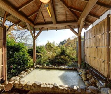 Stone-lined outdoor onsen under wooden pavilion with bamboo screens and garden view in Kanagawa