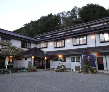 Two-story ryokan exterior with lit entrance, gravel courtyard and wooded hillside backdrop