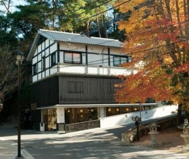 Two-story ryokan exterior with timber facade, orange maple foliage and stone lantern near the entrance in Hiroshima prefecture