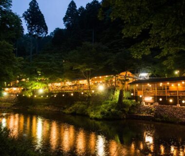 River reflections of lantern-lit wooden ryokan buildings along a forested bank at Momijiya Annex Kyoto