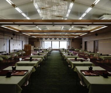 Large ryokan dining hall with rows of low tables set with serving trays, soft lighting and distant windows