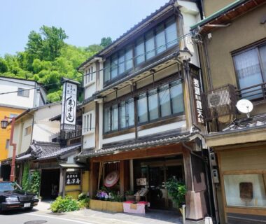 Three-story wooden ryokan building with shoji windows, street entrance and mountain backdrop in Hyōgo, Kansai