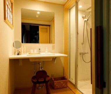 Compact washbasin area with mirror, wooden stool, toiletries and adjacent shower at Motoyu Ishiya, Ishikawa