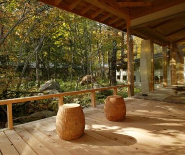 Wooden veranda with two round stools overlooking a forested garden through glass panels