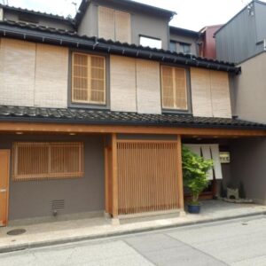 Two-story traditional wooden ryokan entrance with lattice door, bamboo blinds, and potted plants on a narrow street