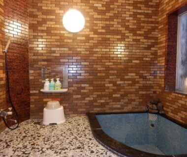 Small tiled indoor bathing room with blue tub, handheld shower, stool, toiletries and window at Murataya in Ishikawa