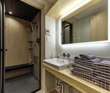 Illuminated mirror, sink and folded towels beside a wood-paneled shower stall with bench in modern ryokan bathroom