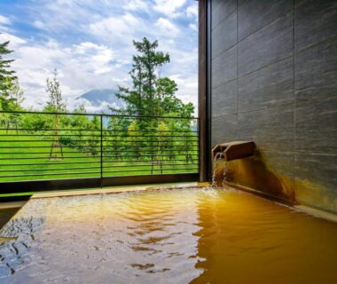 Wooden onsen bath with spout and balcony overlooking lawn and tree-covered hills in Hokkaidō