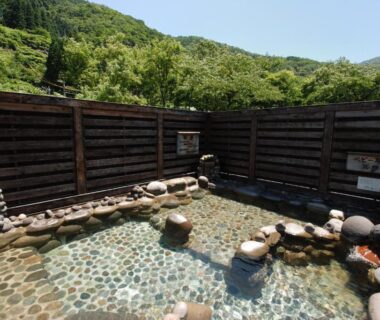 Stone-lined onsen bath with wooden fence and green mountain backdrop