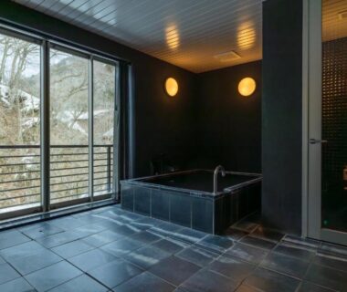 Tiled indoor onsen room with rectangular bath, floor-to-ceiling windows and balcony overlooking wooded hillside at Namari Onsen Iwate