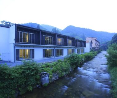 Long white ryokan building beside a shallow stream, rooms with large windows and forested hills beyond in Iwate, Japan