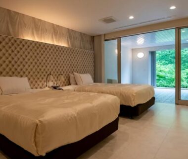 Twin bedroom with padded wall, sliding glass doors to balcony with wooden soaking tub and green hillside view