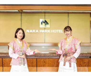 Two reception staff in patterned uniforms gesture at Nara Park Hotel front desk with illuminated hotel sign behind them