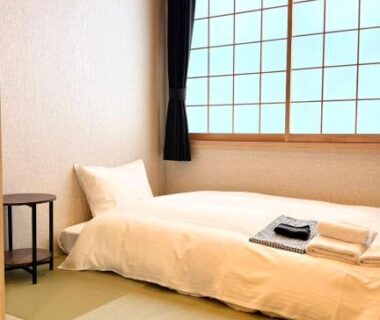 Single bed on tatami with shoji window, folded towels on bedding, small bedside table and wall-mounted air conditioner