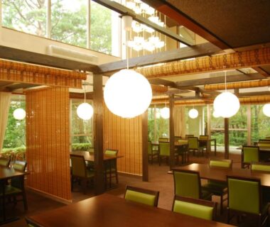 Indoor dining area with wooden partitions, round pendant lights, tables and green chairs overlooking garden