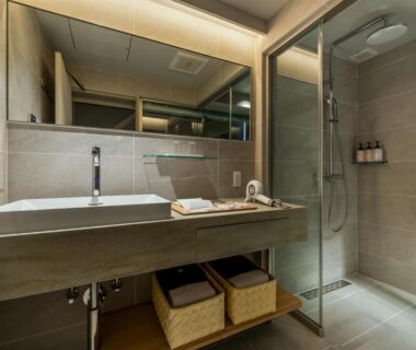 Contemporary bathroom with illuminated mirror, long vanity, glass-enclosed shower, and woven storage baskets at Nazuna Kyoto Tsubaki St. Kyoto