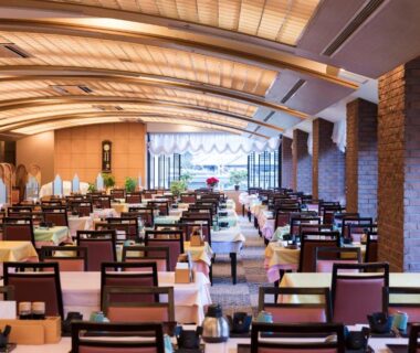 Large ryokan dining hall with rows of set tables, arched ceiling and floor-to-ceiling windows overlooking greenery