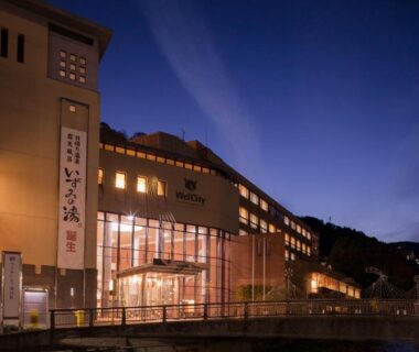 Dusk exterior of New Welcity Yugawara with lit windows along a riverside walkway and hillside backdrop in Shizuoka