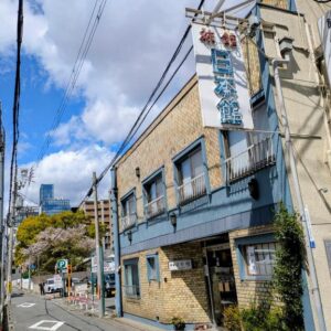 Two-story Nihonkan exterior with signage along a narrow Kyoto street under blue sky