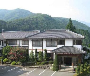Traditional two-story ryokan exterior with tiled roof, landscaped entrance and parking, mountains behind in Gunma, Kantō, Japan