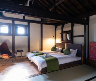 Twin low beds with green runners on tatami mats under exposed wooden beams, butterfly chair and decorative screen