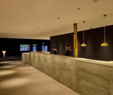 Long concrete reception counter with staff, pendant lights and wooden pillars in minimalist ryokan lobby