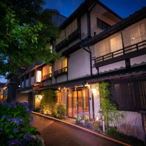 Night view of lit traditional ryokan entrance with wooden facade, shoji screens, lanterns and flowered pathway in Kyoto