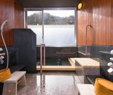 Wood-paneled indoor onsen with washing stations, stools, wooden buckets and window overlooking hills at Oishiya, Mie