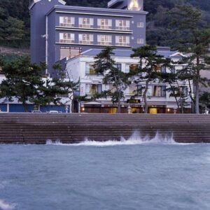 Multi-story building of Oishiya on seawall with waves, shoreline pines and forested hillside in Mie, Kansai