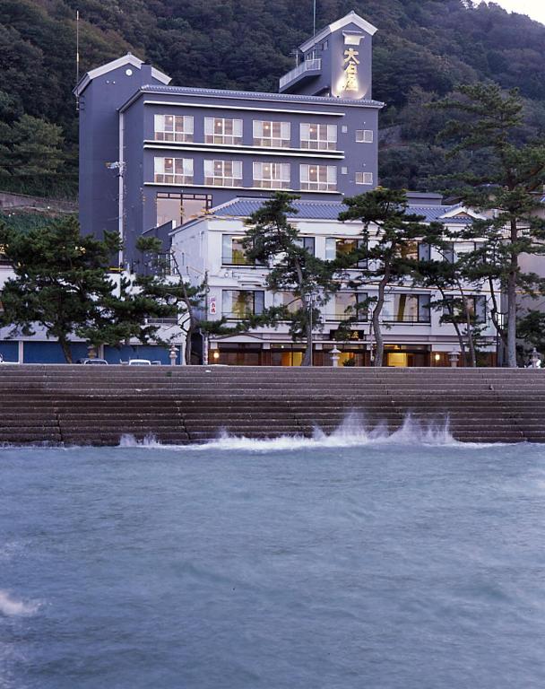 Seaside facade at Oishiya Mie Multi-story building of Oishiya on seawall with waves, shoreline pines and forested hillside in Mie, Kansai