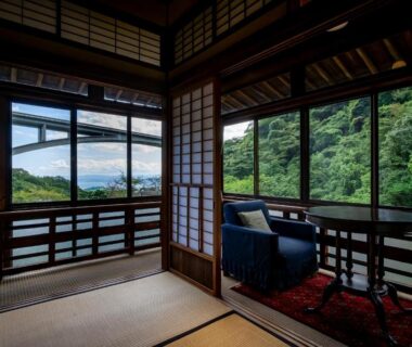 Tatami room with shoji screens and seating overlooking green hillside and distant water in Ōita
