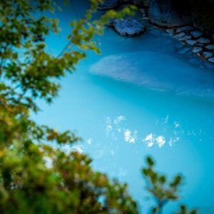 Milky turquoise onsen water with stone edging and lush greenery at Okamotoya in Ōita, Kyūshū