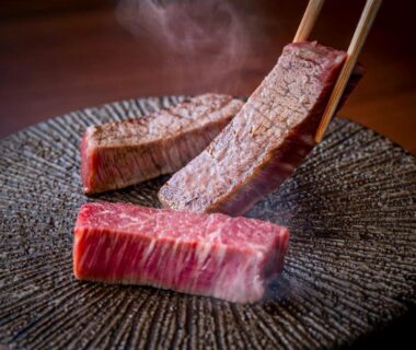Seared beef on dark ceramic plate with tongs, steam rising, dining at Okamotoya in Ōita, Kyūshū