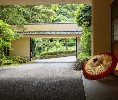 Sheltered ryokan entrance with traditional umbrella, shoji screen and gravel driveway opening onto a lush garden in Kanagawa