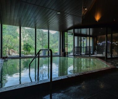 Indoor onsen pool with handrail and floor-to-ceiling windows overlooking garden at Okuhita Onsen Umehibiki, Ōita