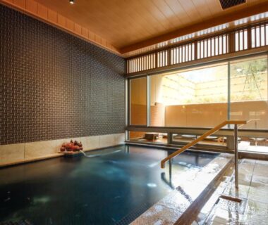 Indoor onsen bath with wooden ceiling, tiled walls, handrails and windows overlooking bamboo screen