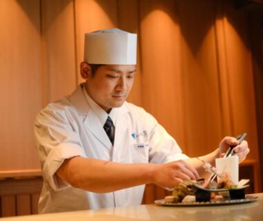 Chef in white uniform plating dishes behind a wooden counter in Onomichi Hansei dining area, Hiroshima
