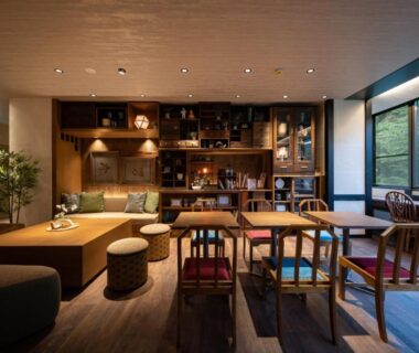 Wooden lounge with low seating, stools, bookshelf, and large window showing lush greenery at Onsen Guest House Tsutaya Kanagawa