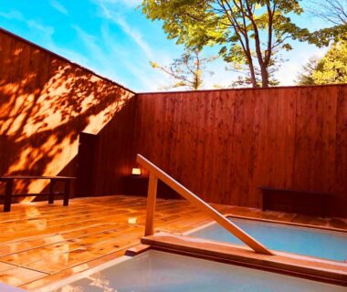 Walled wooden outdoor onsen deck with two baths, handrail and bench under blue sky