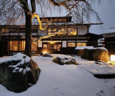 Snow-covered courtyard with lanterns and rocks, warm lit wooden facade at Onsen and Garden Nana, Ishikawa, Chūbu