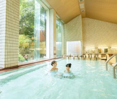 Guests bathing in large indoor onsen with tall windows and tiled walls at Ooedo Onsen Monogatari Awazu Grand Hotel Ishikawa