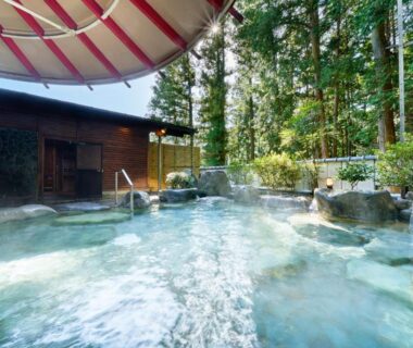 Steamy outdoor stone onsen bath with wooden building and tall cedar trees at Ooedo Onsen Monogatari Gero Bekkan Gifu